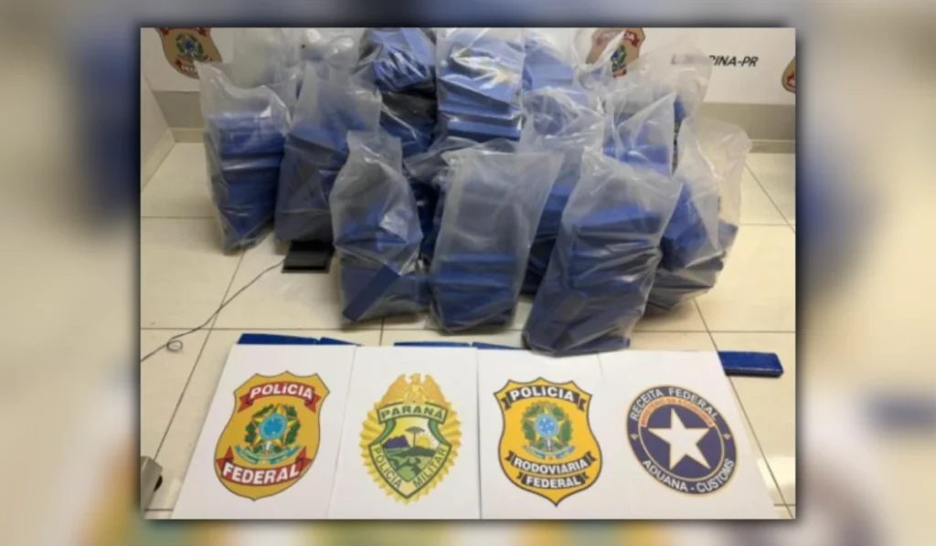 Centenas de tabletes azuis de maconha empilhados no chão com os brasões da Polícia Federal, Polícia Militar do Paraná, Polícia Rodoviária Federal e Receita Federal posicionados à frente durante apreensão de maconha em Rolândia.